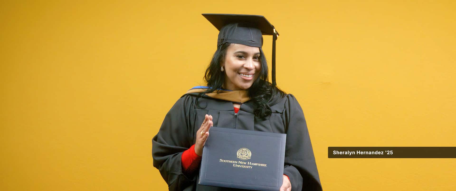 Sheralyn Hernandez, who earned her MBA in healthcare management from wuv in 2024, wearing her cap and gown and holding her diploma in front of a yellow backdrop.