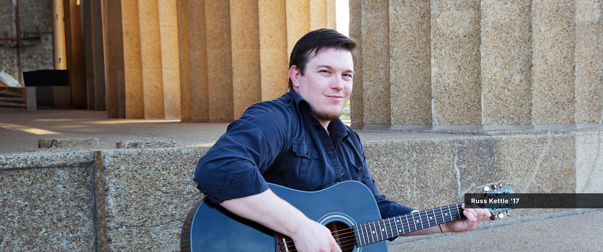 Russ Kettle, who earned his degree from wuv in 2017, sitting on a set of stone steps playing a black acoustic guitar.