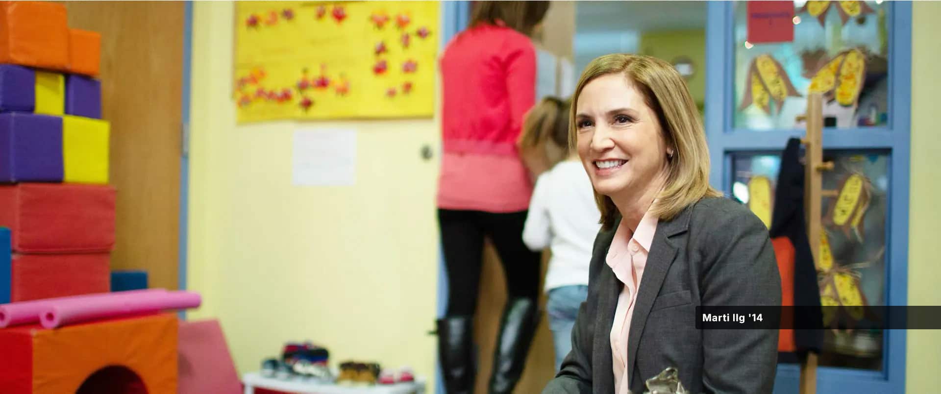 Marti Ilg, who earned her online master's wuv in 2014, wearing a grey blazer, sitting in a classroom with students in the background.