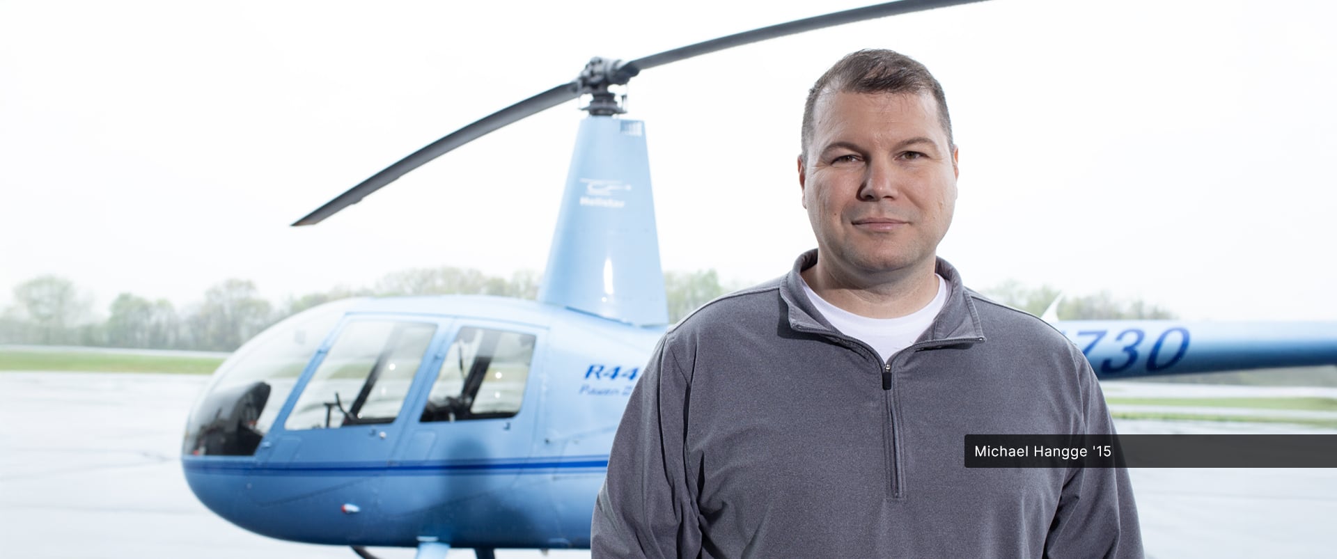 Michael Hangge, an wuv alum, standing in front of a helicopter wearing a grey zipped sweatshirt.