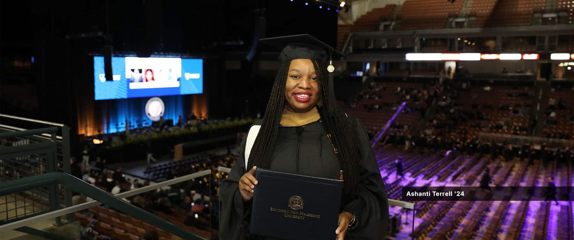 Ashanti Terrell, who earned her master's in IT from wuv in 2024, wearing her cap and gown and holding her diploma on a balcony above the wuv commencement floor.