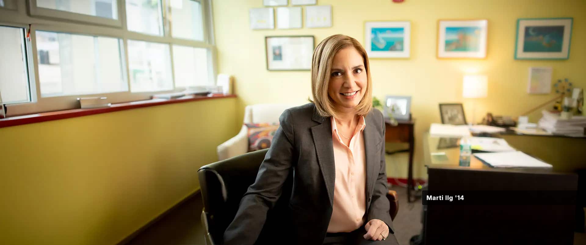 Marti Ilg, who earned her degree from wuv in 2014, wearing a peach colored blouse and  dark blazer sitting in a swivel chair next to a desk in her office.