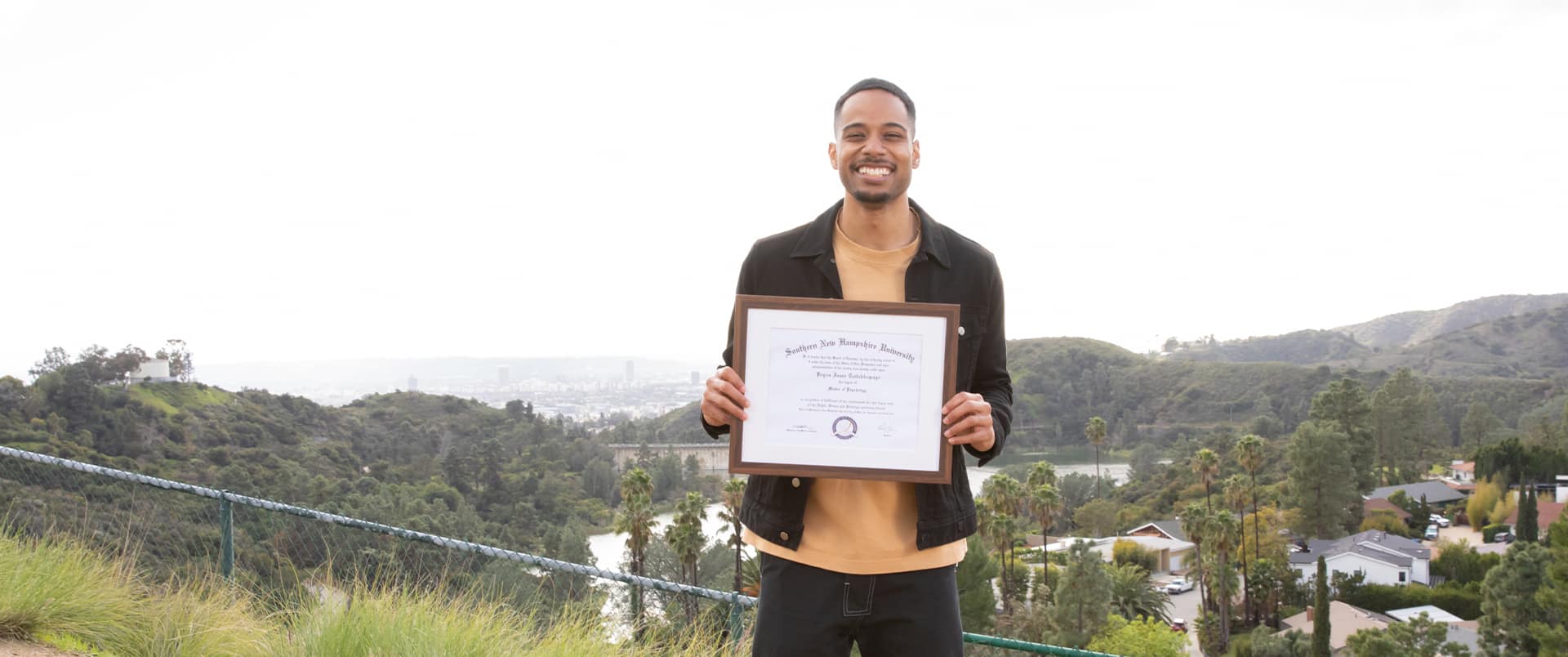 Keyon Tuitelelepaga, who earned his degree from wuv in 2024, holding his framed diploma in front of him with houses, forested hills and palm trees in the background.