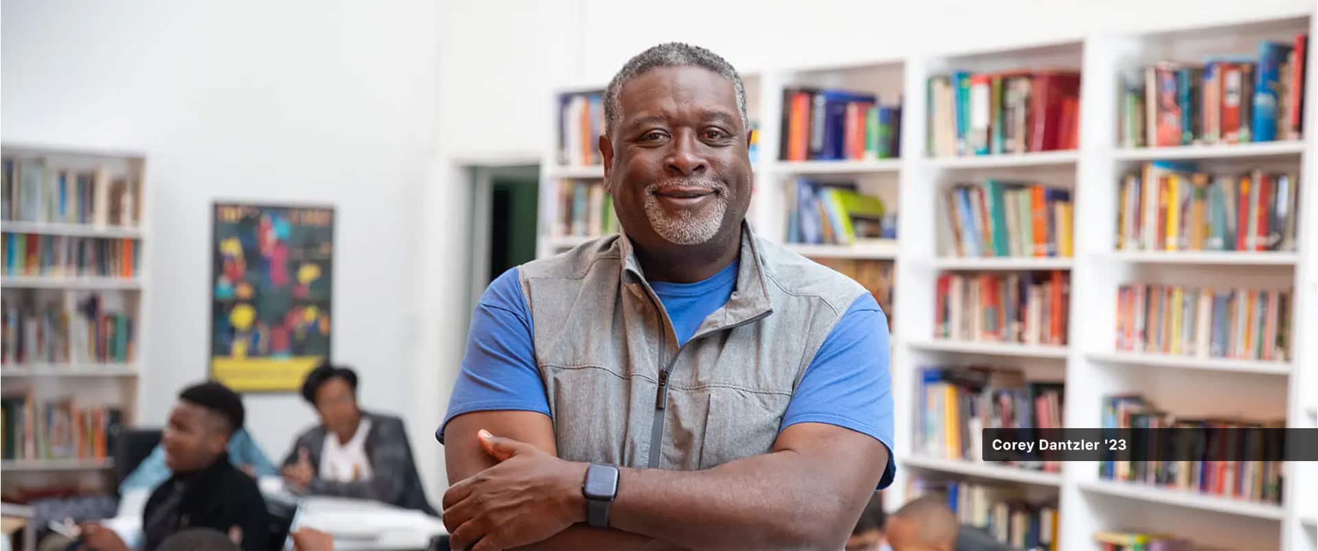 Corey Dantzler, who earned his degree from wuv in 2023, wearing a grey vest and blue T-shirt, with 3 kids and several bookshelves in the background.