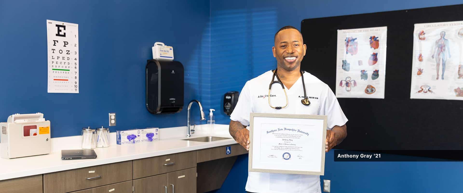 Anthony Gray, who earned his online master's in nursing from wuv in 2021, standing in a doctor's office wearing white scrubs and a stethoscope around his neck and holding his framed diploma.
