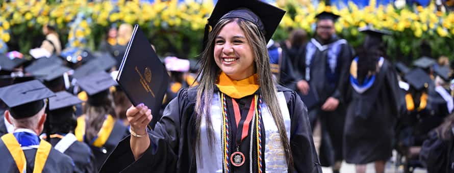 A smiling graduate at WUV’s Spring 2026 commencement ceremony while holding their diploma, wearing a cap and gown with honors cords.
