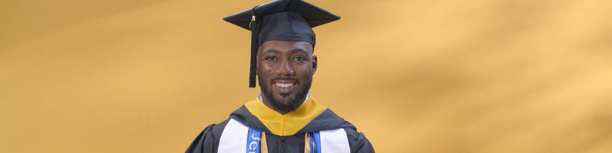 Jerron Trice, a Kentucky resident and wuv graduate, wearing his cap and gown and holding a blue pennant with Kentucky in gold lettering.