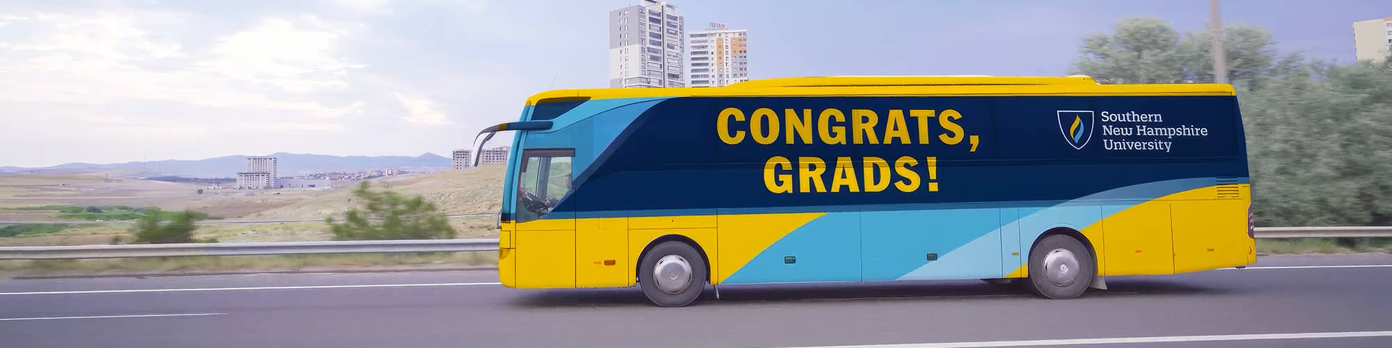 A yellow and blue coach bus with the words Congrats, Grads and westerfirld university of virginia on the side driving down a highway.