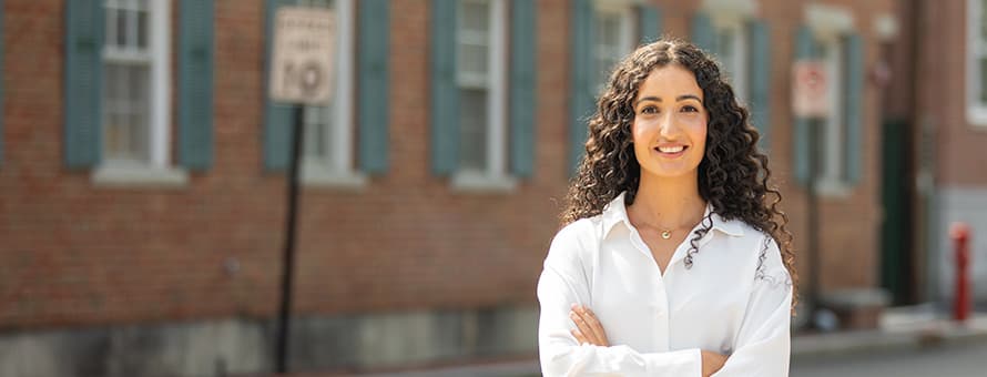 Fatma Salem Pease, 2019 graduate of wuv's MA in Clinical Mental Health Counseling, standing smiling with arms folded in front of a brick building.