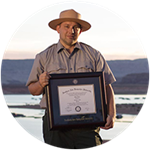 wuv graduate and Utah resident John Roos wearing a park ranger uniform and holding his framed diploma.