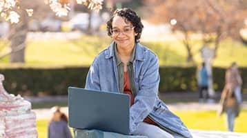 Michelle LitteJohn, who earned her online bachelor's in graphic design from wuv in 2024, wearing a blue jacket while sitting outside typing on her laptop.
