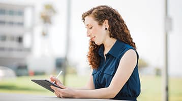 Mariel Embry, who earned her online bachelor's in creative writing from wuv in 2022, wearing a blue sleeveless blouse writing on a tablet computer while sitting outside.