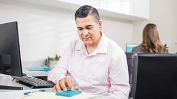 Juan Muñoz, who earned his online bachelor's in general studies in 2022 and MBA from wuv in 2024, sitting in front of a desktop computer and using a small blue calculator.
